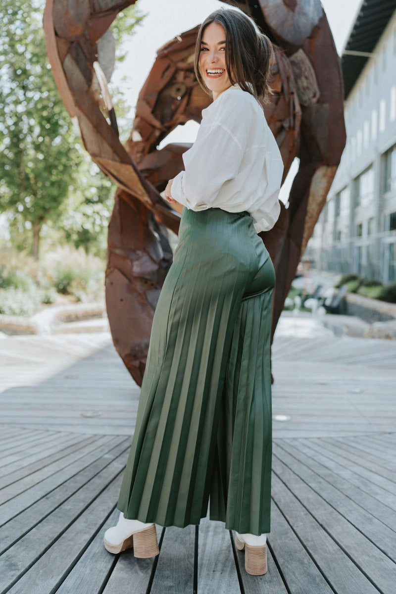 Back view of model wearing the Make You Mine Pants, which feature a hunter green faux-leather material, a pleated design, an elastic waist, and a wide leg fit. Worn with white top.