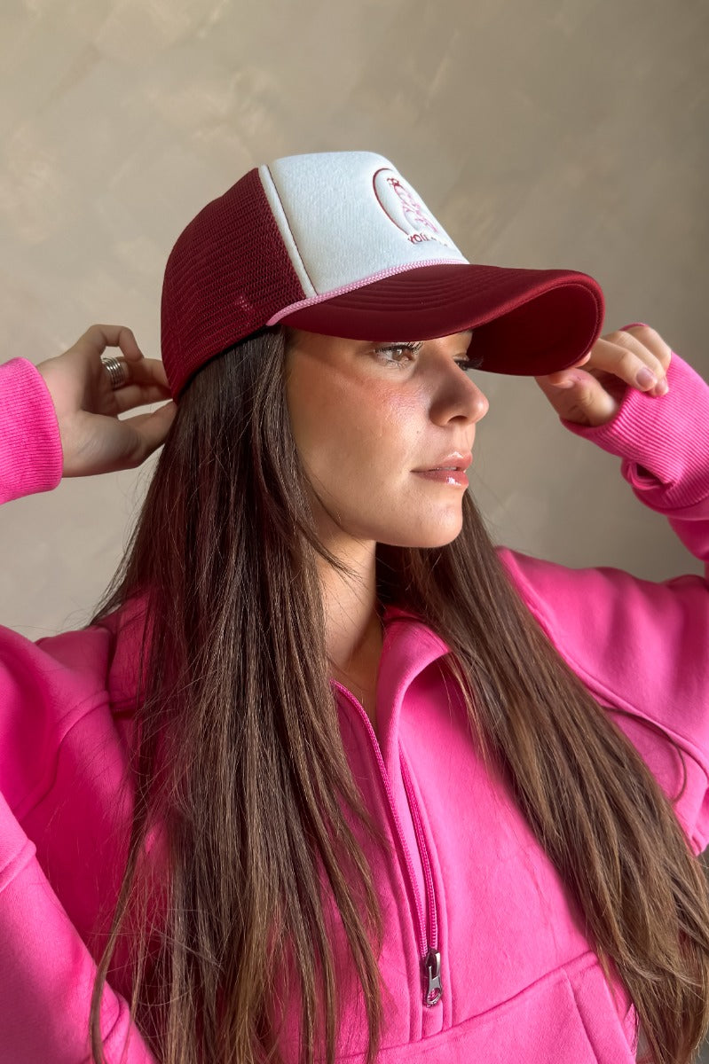 Side view of model wearing the Breast Cancer Gear: You Can Do It Trucker Hat that has a burgundy bill and back, a snapback, pink trim, and a cream front with an embroidered pink and burgundy graphic. 