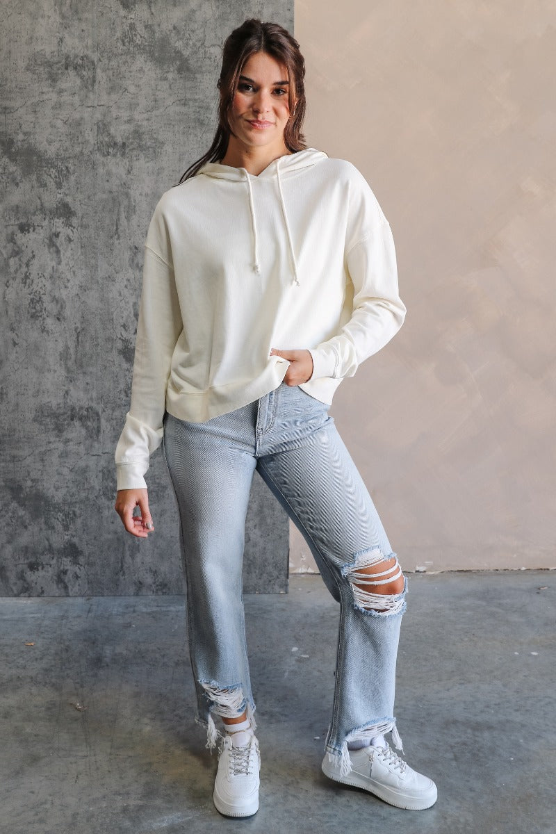 Full body view of model wearing the Chloe Cream Hoodie Long Sleeve Sweatshirt which features cream cotton fabric, a thick hem, a high neckline with a hood attached and drawstring ties, a dropped shoulders, and long sleeves with cuffs.