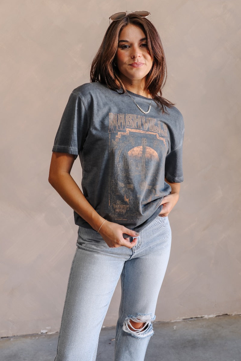 Front view of model wearing the Nashville Charcoal Grey Short Sleeve Graphic Tee which features dark grey washed cotton fabric, round neckline and short sleeves. Graphic says "Nashville" with a guitar and music city scene graphic in light grey, rust, and 