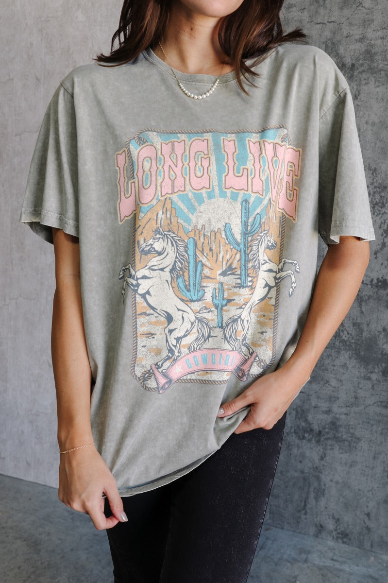 Front view of model wearing the Long Live Cowgirls Grey Graphic Tee which features washed grey cotton fabric, round neckline and short sleeves. Graphic says "Long Live Cowgirls" with two horses and desert scene graphic in pink, blue, dark yellow and off w