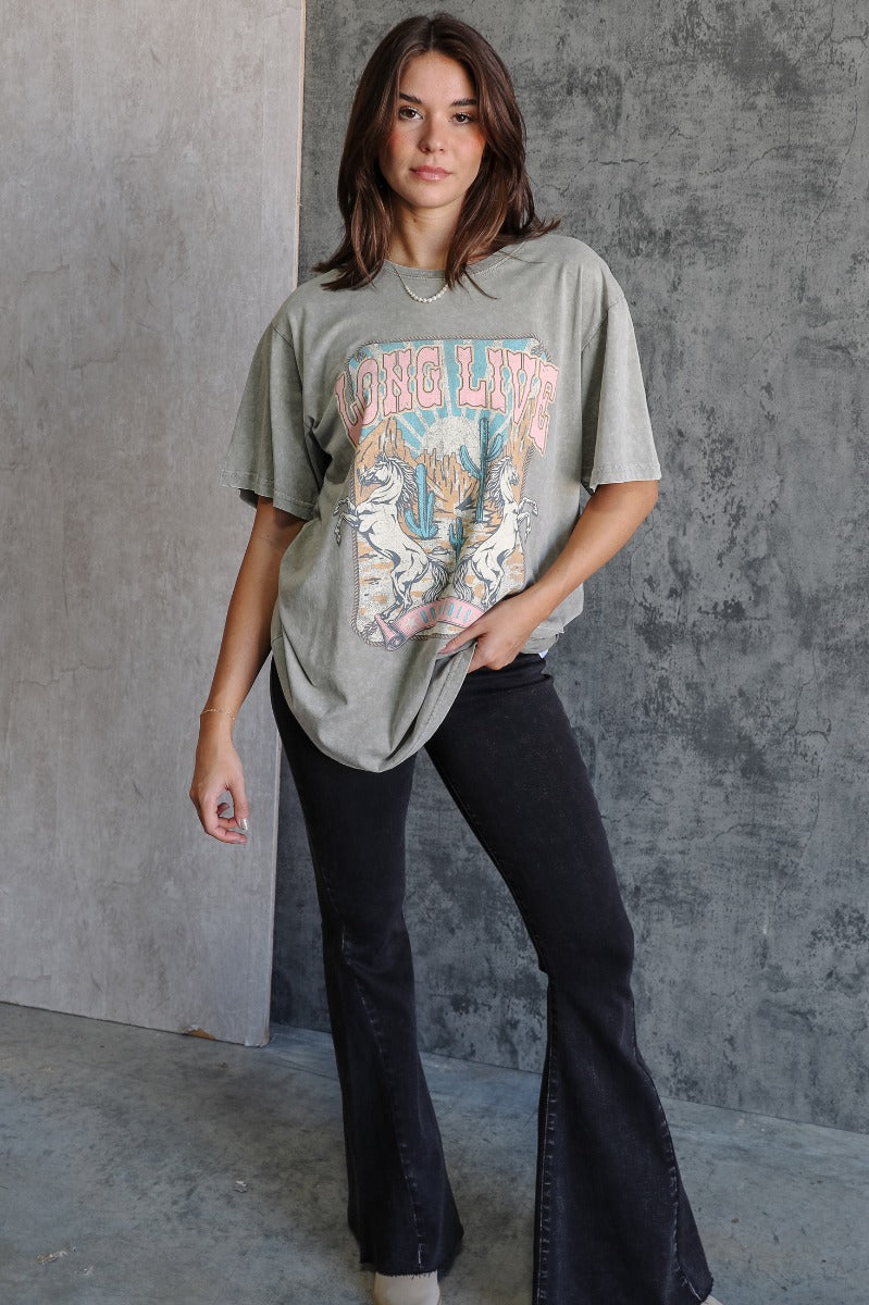 Full body view of model wearing the Long Live Cowgirls Grey Graphic Tee which features washed grey cotton fabric, round neckline and short sleeves. Graphic says "Long Live Cowgirls" with two horses and desert scene graphic in pink, blue, dark yellow and o