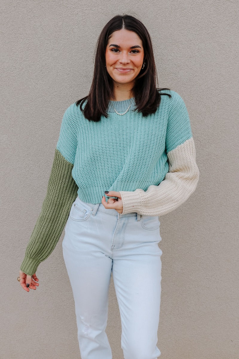 Front view of model wearing the Celeste Sage Multi Long Sleeve Sweater which features sage, light blue and cream cable knit fabric, a colorblock pattern, a cropped waist, ribbed trim, a round neckline, dropped shoulders, and long balloon sleeves with cuff