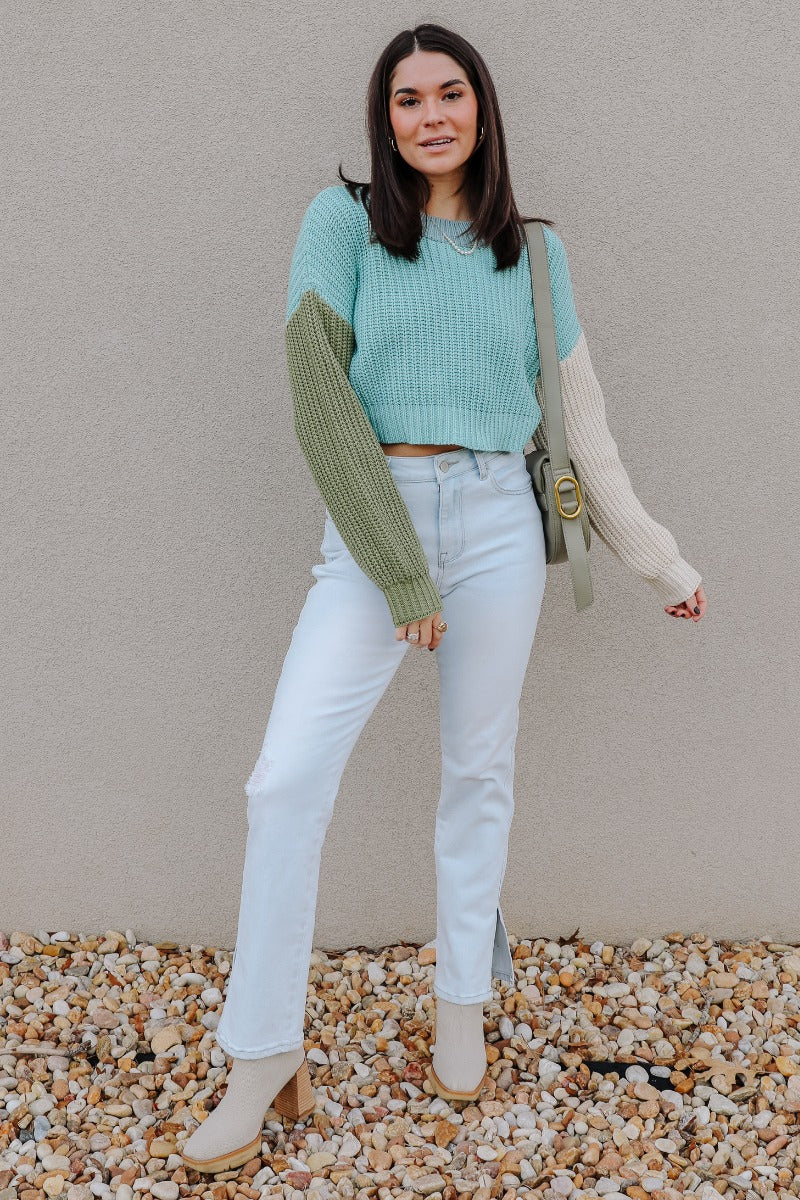 Full body view of model wearing the Celeste Sage Multi Long Sleeve Sweater which features sage, light blue and cream cable knit fabric, a colorblock pattern, a cropped waist, ribbed trim, a round neckline, dropped shoulders, and long balloon sleeves with 