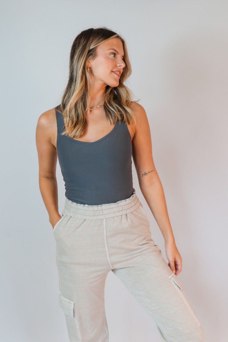 Front view of model wearing the Avery Charcoal Grey Sleeveless Bodysuit which features charcoal grey knit fabric, a scooped neckline, thick straps, and a thong bottom with snap closures.