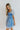 Side view of model wearing the Forget Me Not Babydoll Dress that has blue and light blue organza fabric with a floral pattern, a mini-length hem, a square neck, a back zipper with a hook closure, and short puff sleeves