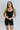 Front view of model wearing the Can You Keep Up Bodysuit that features black stretch fabric, a round neckline, adjustable thick straps, and fitted shorts.