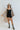 Full body front view of model wearing the Can You Keep Up Bodysuit that features black stretch fabric, a round neckline, adjustable thick straps, and fitted shorts. Worn with shirt dress.