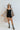 Full body front view of model wearing the Can You Keep Up Bodysuit that features black stretch fabric, a round neckline, adjustable thick straps, and fitted shorts. Worn with shirt dress.