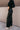 Side view of model wearing the Alexis Black Faux Leather Pants which features black faux leather, front zipper with button closure, two front pockets, two back pockets, belt loops and wide pant legs.