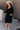 Full body view of model wearing the Wrenley Black Long Sleeve Midi Dress which features black gauze fabric, a tiered body, midi-length hem, long balloon sleeves with elastic trim, side pockets, a v neckline, and a smocked back.