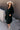 Full body view of model wearing the Wrenley Black Long Sleeve Midi Dress which features black gauze fabric, a tiered body, midi-length hem, long balloon sleeves with elastic trim, side pockets, a v neckline, and a smocked back.