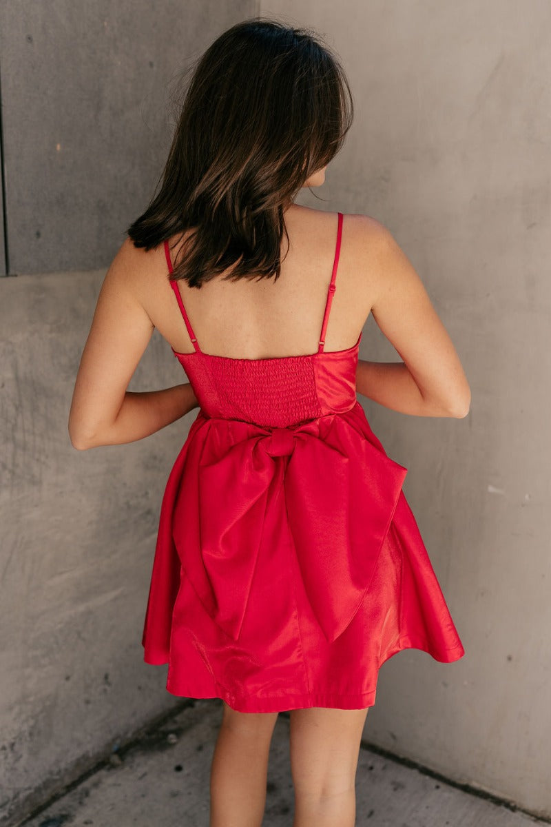 Back view of model wearing the Avery Red Bow Mini Dress which features crimson red sheen fabric, red lining, mini length, two front pockets, sweetheart neckline, adjustable straps, sleeveless, monochromatic side zipper with hook closure, smocked back and 