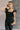 Upper front view of model wearing the Sabrina Black Denim Sleeveless Jumpsuit that has washed black denim cotton fabric, pockets, an elastic waist, a zip-up square neck, ruffle straps, and straight legs.