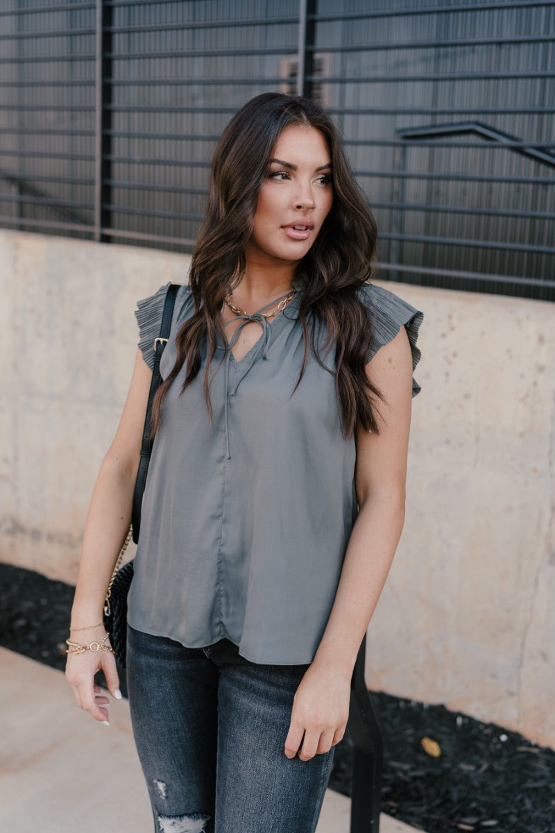 Front view of model wearing the Daniela Charcoal Satin Pleated Top that has charcoal grey satin fabric, a v-neck with ruffle details, an adjustable tie, and short sleeves with pleated details.