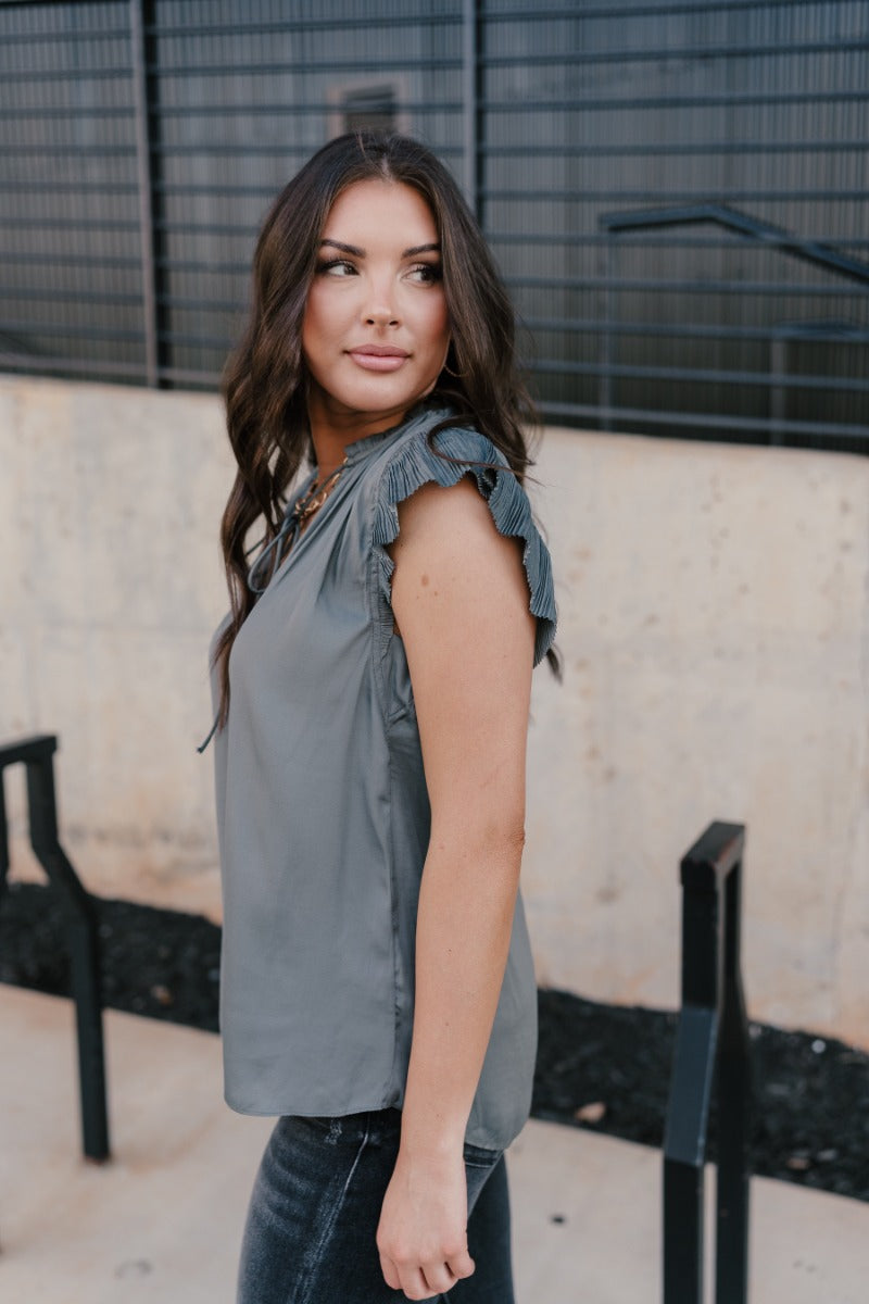 Side view of model wearing the Daniela Charcoal Satin Pleated Top that has charcoal grey satin fabric, a v-neck with ruffle details, an adjustable tie, and short sleeves with pleated details.