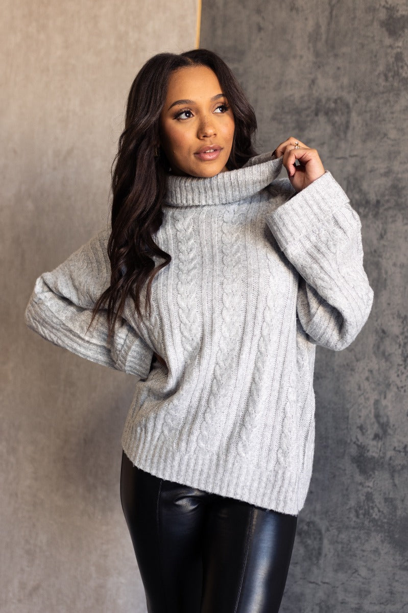 Front view of model wearing the Palmer Grey Knit Turtleneck Long Sleeve Sweater which features light grey knit fabric, a monochrome cable knit design, ribbed trim, a turtleneck neckline, dropped shoulders, and long sleeves with ribbed cuffs.