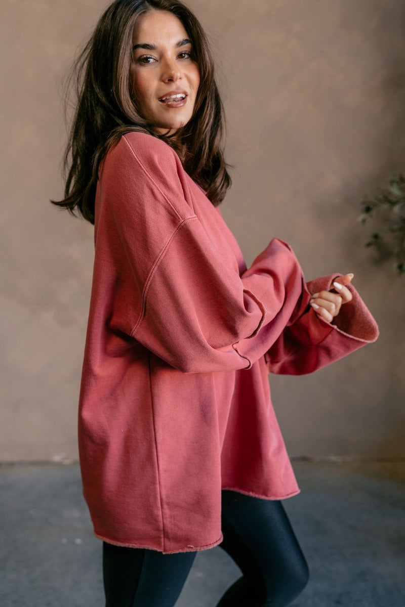 Front view of model wearing the Astro Brick Red Long Sleeve Sweatshirt which features bricked red knit fabric, a raw hem, a ribbed round neckline, dropped shoulders, and long sleeves with raw ruffle details.