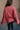 Back view of model wearing the Astro Brick Red Long Sleeve Sweatshirt which features bricked red knit fabric, a raw hem, a ribbed round neckline, dropped shoulders, and long sleeves with raw ruffle details.