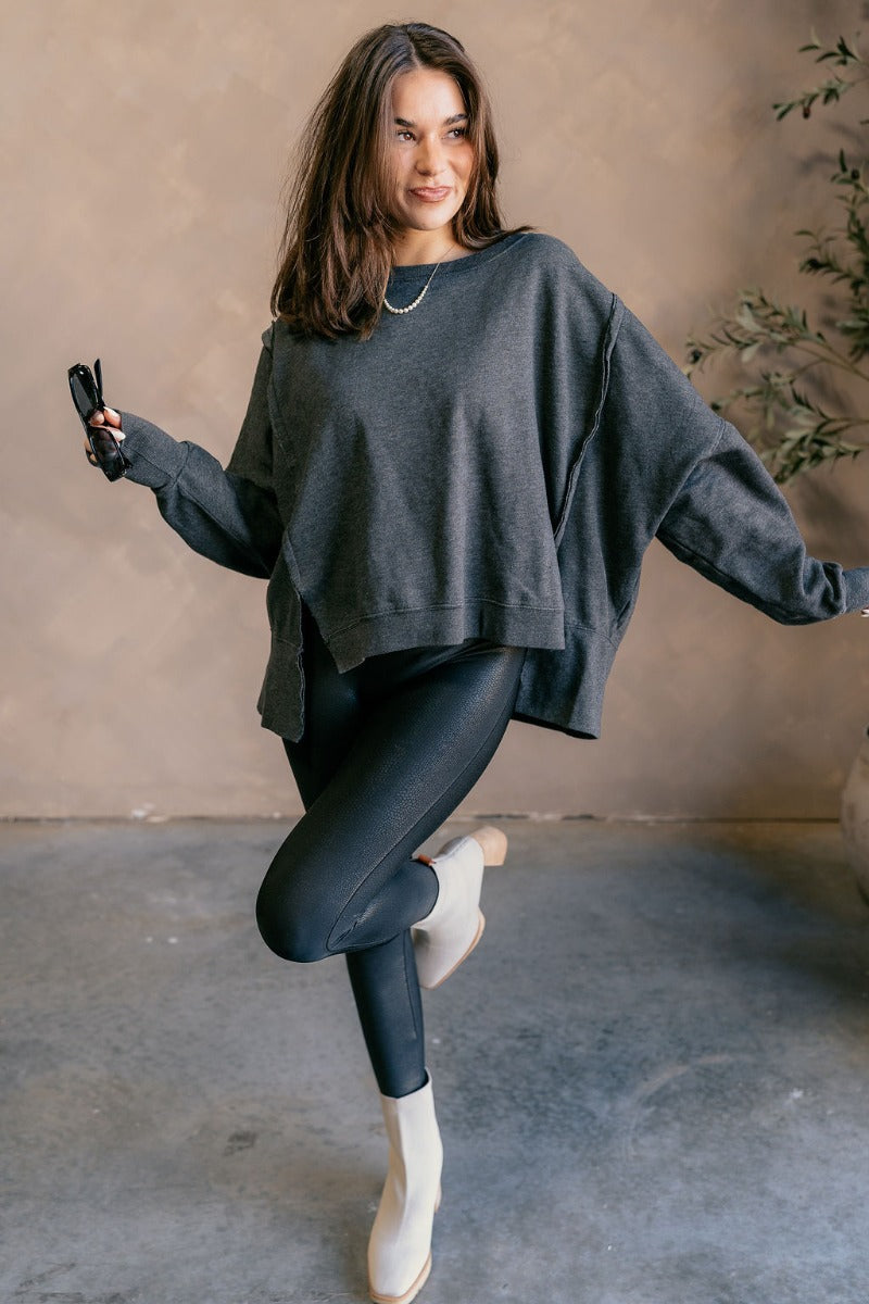 Full body view of model wearing the Lyla Washed Black Long Sleeve Sweatshirt which features washed black knit fabric, a ribbed hem with slight slits on each side, raw hem details, a round ribbed neckline, dropped shoulders and long balloon sleeves with ri