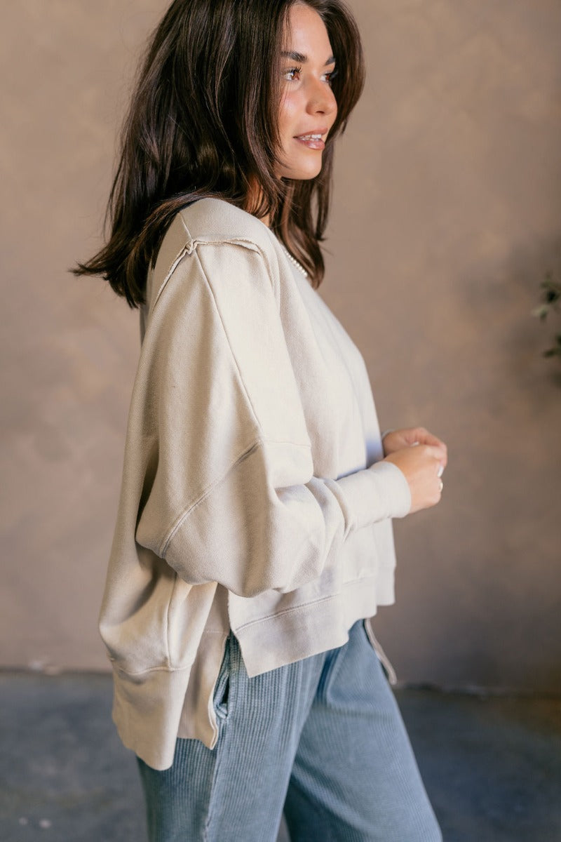 Side view of model wearing the Lyla Cream Long Sleeve Sweatshirt which features washed black knit fabric, a ribbed hem with slight slits on each side, raw hem details, a round ribbed neckline, dropped shoulders and long balloon sleeves with ribbed cuffs.