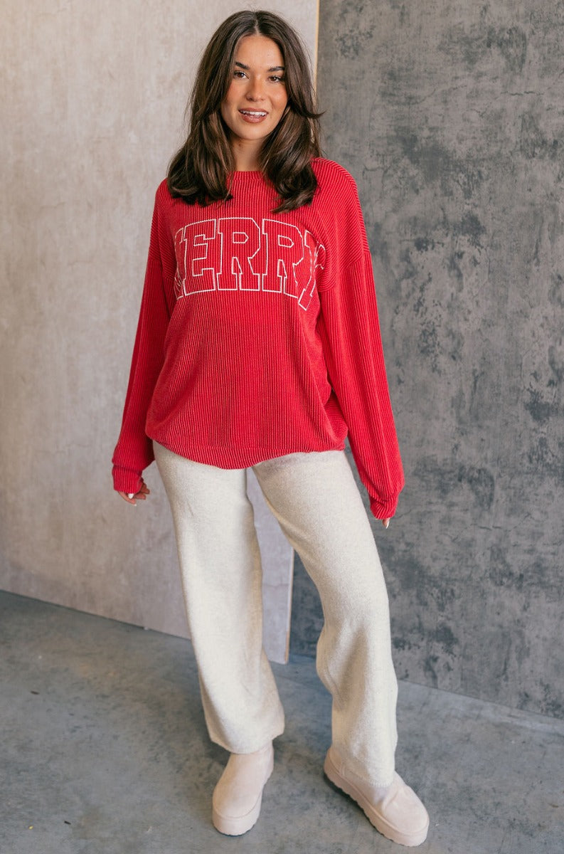 Full body view of model wearing the Merry Red Ribbed Long Sleeve Sweater which features red and light red ribbed fabric, thick hem, a round neckline, a graphic that says "MERRY" in white outline, dropped shoulders, and long sleeves with ribbed cuffs.