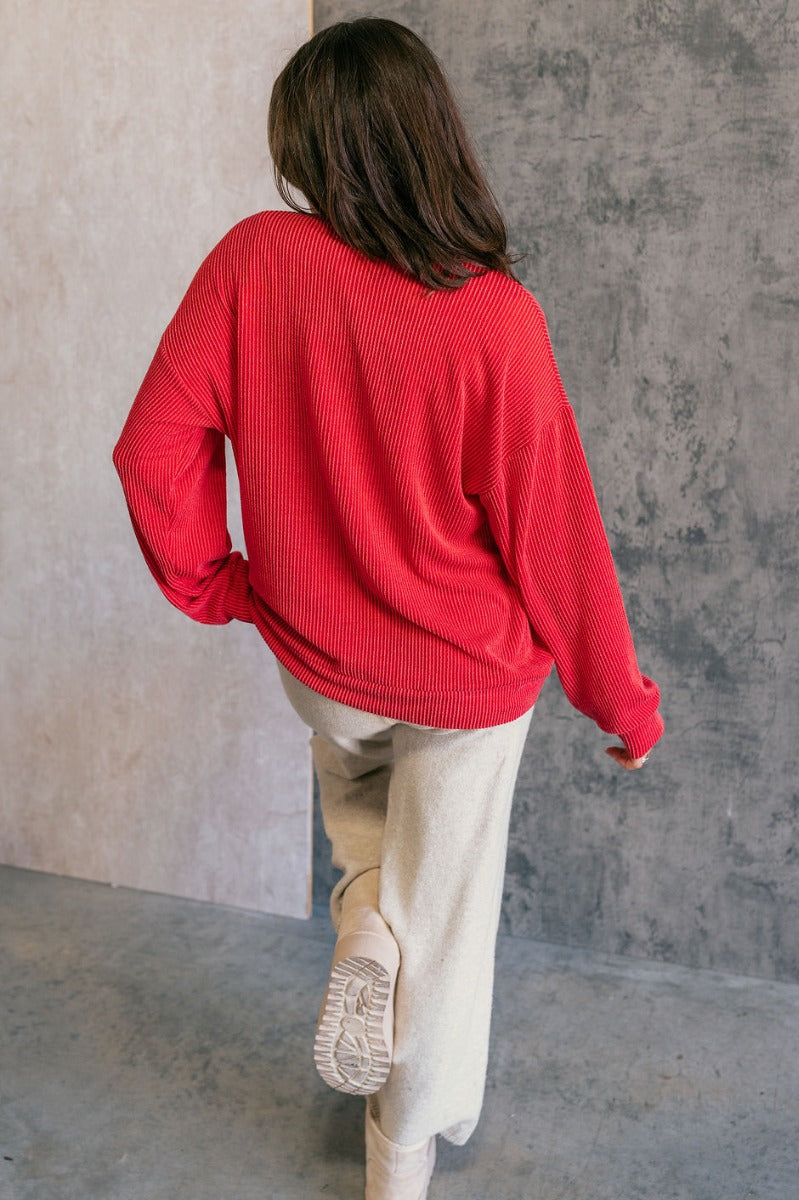 Full body back view of model wearing the Merry Red Ribbed Long Sleeve Sweater which features red and light red ribbed fabric, thick hem, a round neckline, a graphic that says "MERRY" in white outline, dropped shoulders, and long sleeves with ribbed cuffs.