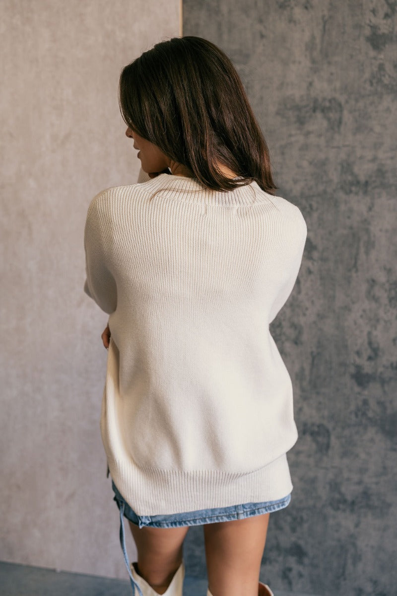 Back view of model wearing the Milena Cream Long Sleeve Sweater that has cream knit fabric, ribbed hem, ribbed high neckline, dropped shoulders and long sleeves with ribbed cuffs. Shown untucked.