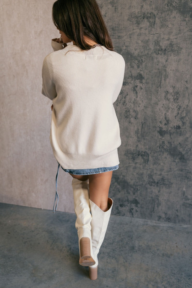 Full body back view of model wearing the Milena Cream Long Sleeve Sweater that has cream knit fabric, ribbed hem, ribbed high neckline, dropped shoulders and long sleeves with ribbed cuffs. Shown untucked.