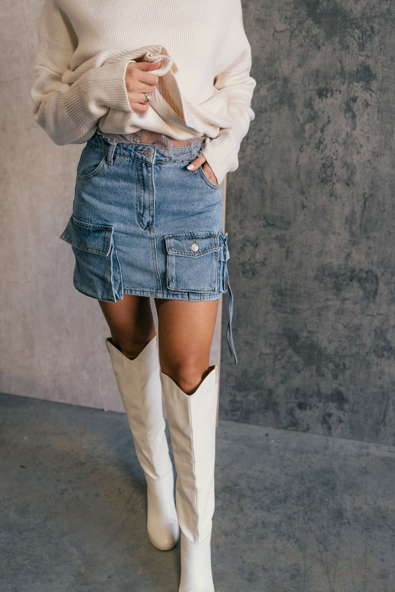 Front view of model wearing the Samira Blue Denim Mini Cargo Skirt that has blue denim fabric, mini length, pockets, belt loops and a front zipper with a button closure.