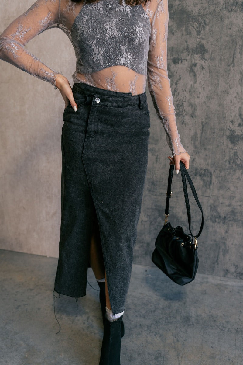 Front view of model wearing the Harlow Black Denim Front Slit Midi Skirt that has washed black denim fabric, a raw hem, midi length, a front slit, a slanted front zipper, pockets, and belt loops.