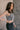 Side view of model wearing the Nadia Grey Lace Long Sleeve Top that has grey sheer fabric, lettuce hem details, a lace floral design, a high neckline, and long sleeves. Worn tucked into black skirt over black tank.
