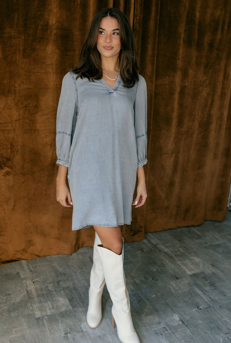 Full body view of model wearing the Naomi Denim Blue Half Sleeve Mini Dress which features washed blue denim fabric, mini length, slight slits on each side, pockets on each side, a round neckline with a v cutout, and half balloon sleeves.