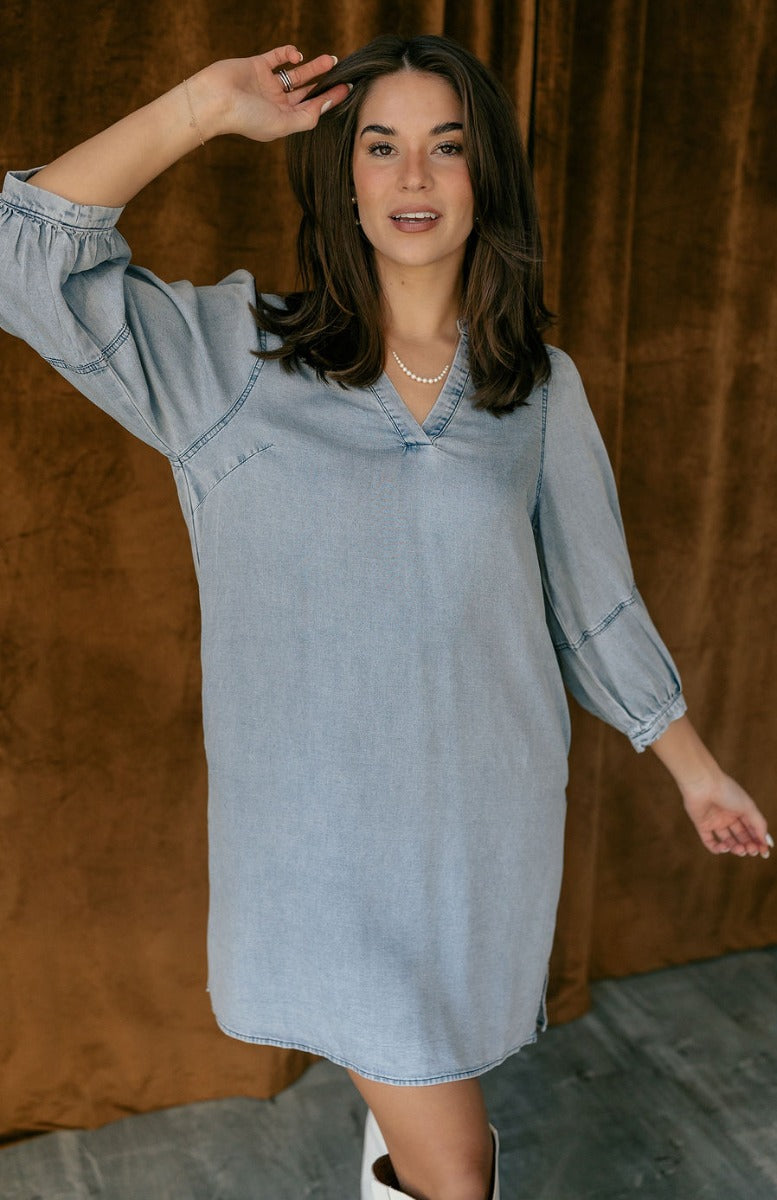 Front view of model wearing the Naomi Denim Blue Half Sleeve Mini Dress which features washed blue denim fabric, mini length, slight slits on each side, pockets on each side, a round neckline with a v cutout, and half balloon sleeves.