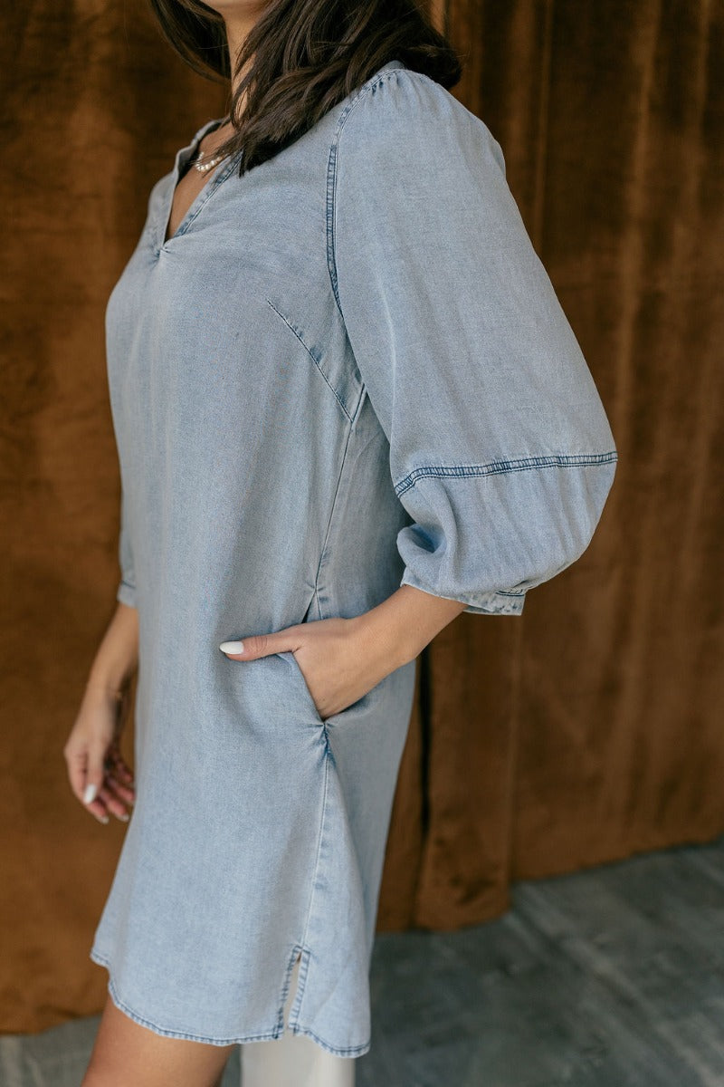 Side view of model wearing the Naomi Denim Blue Half Sleeve Mini Dress which features washed blue denim fabric, mini length, slight slits on each side, pockets on each side, a round neckline with a v cutout, and half balloon sleeves.