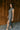 Full body side view of model wearing the Alianna Silver Shimmer Cowl Neck Midi Dress that has grey and silver shimmer fabric, midi length, ruched side details, a slit on the side, a slight cowl high neckline, and a sleeveless design.