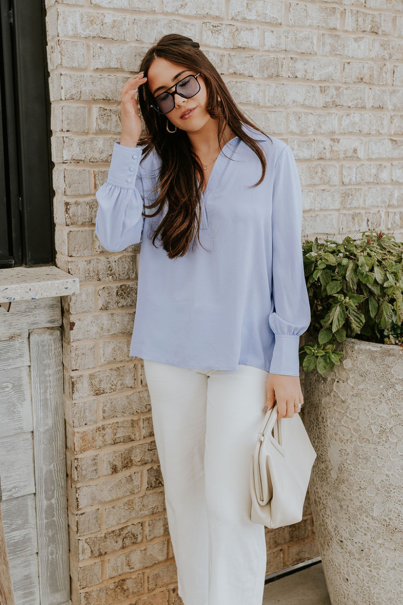Full body view of model wearing the Chloe Light Blue Buttoned Long Sleeve Top which features light blue light weight fabric, small slits on each side, collared v-neckline and long sleeves with monochrome buttoned cuffs.