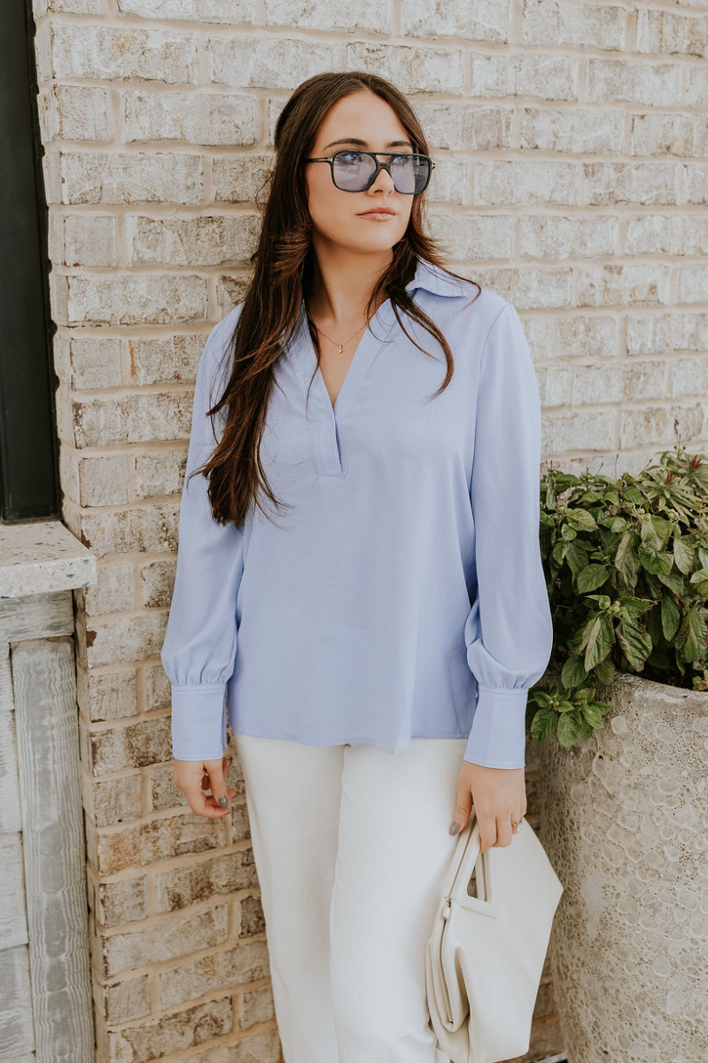 Front view of model wearing the Chloe Light Blue Buttoned Long Sleeve Top which features light blue light weight fabric, small slits on each side, collared v-neckline and long sleeves with monochrome buttoned cuffs.