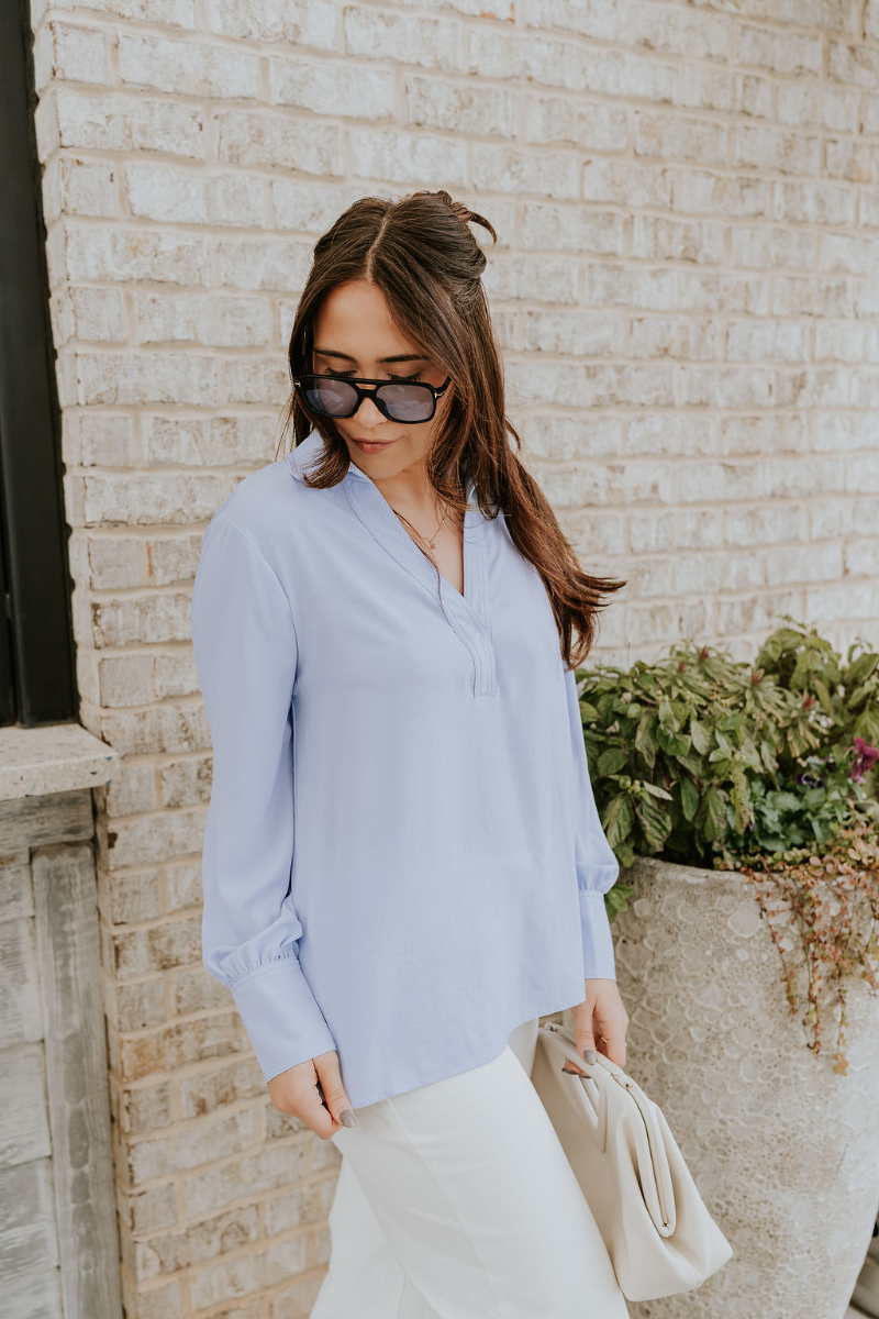 Side view of model wearing the Chloe Light Blue Buttoned Long Sleeve Top which features light blue light weight fabric, small slits on each side, collared v-neckline and long sleeves with monochrome buttoned cuffs.