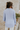 Back view of model wearing the Chloe Light Blue Buttoned Long Sleeve Top which features light blue light weight fabric, small slits on each side, collared v-neckline and long sleeves with monochrome buttoned cuffs.
