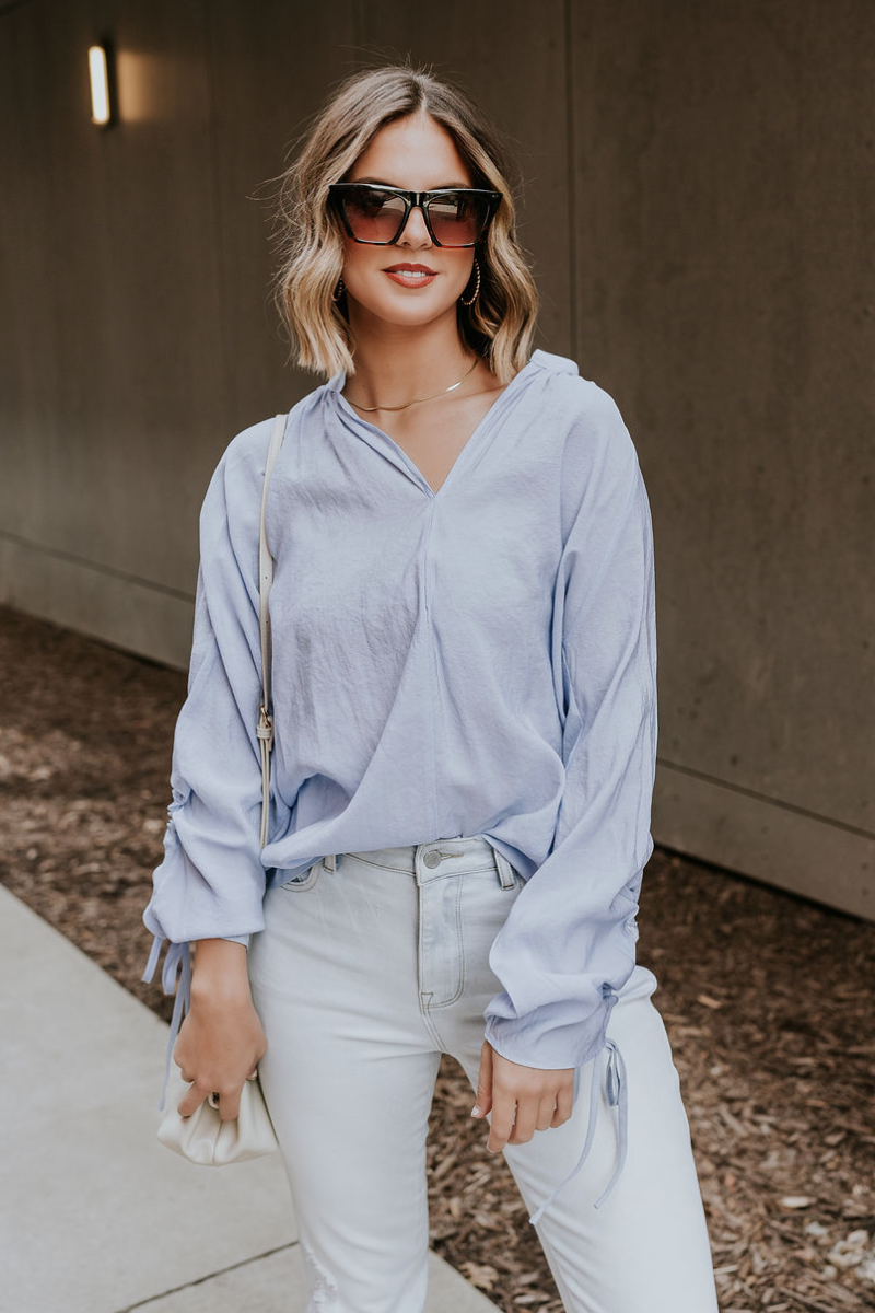 Front view of model wearing the Selene Light Blue Long Sleeve Top which features light blue light weight fabric, v-neckline with collar and long sleeves with an open key hole and cinched details.