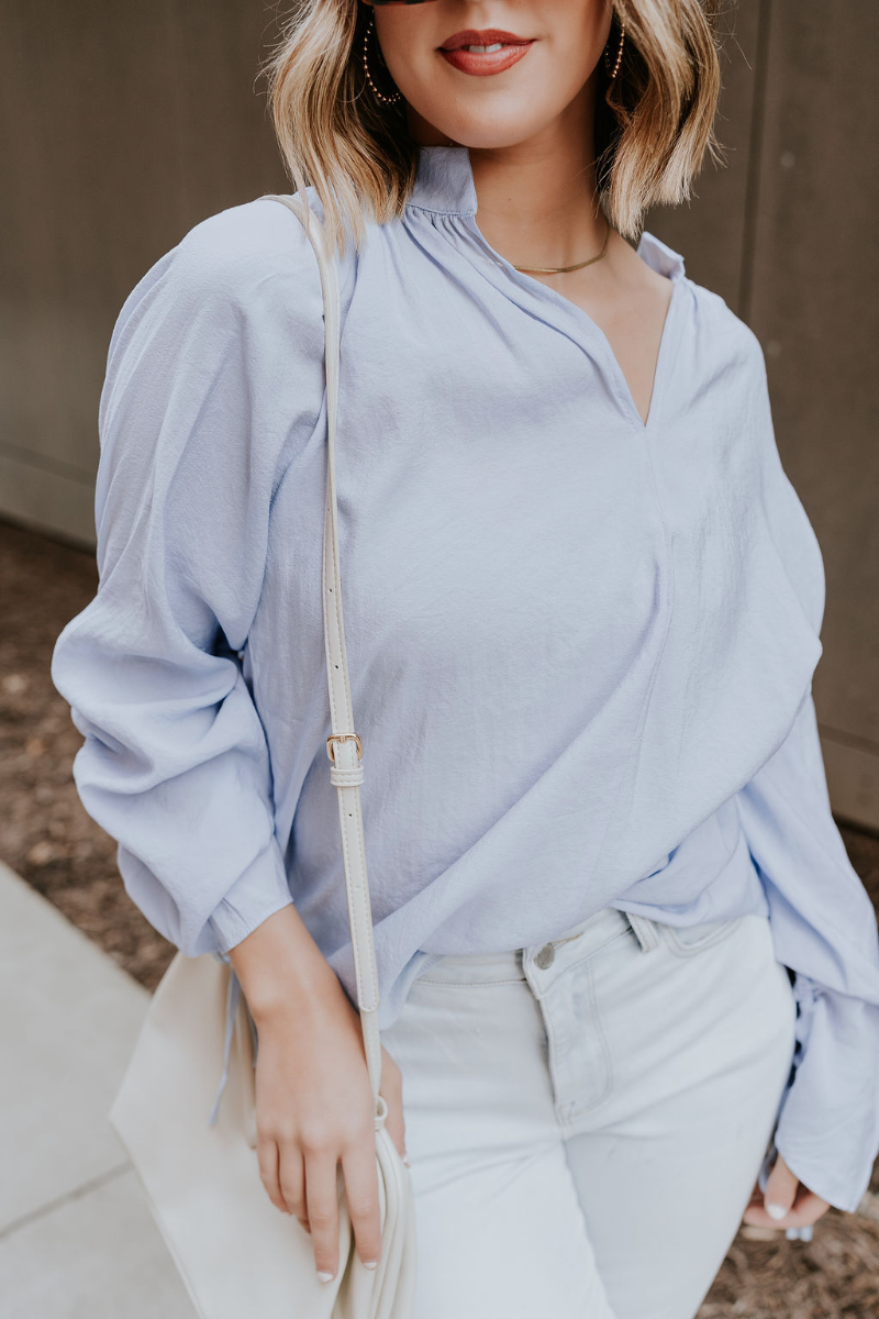 Close up view of model wearing the Selene Light Blue Long Sleeve Top which features light blue light weight fabric, v-neckline with collar and long sleeves with an open key hole and cinched details.