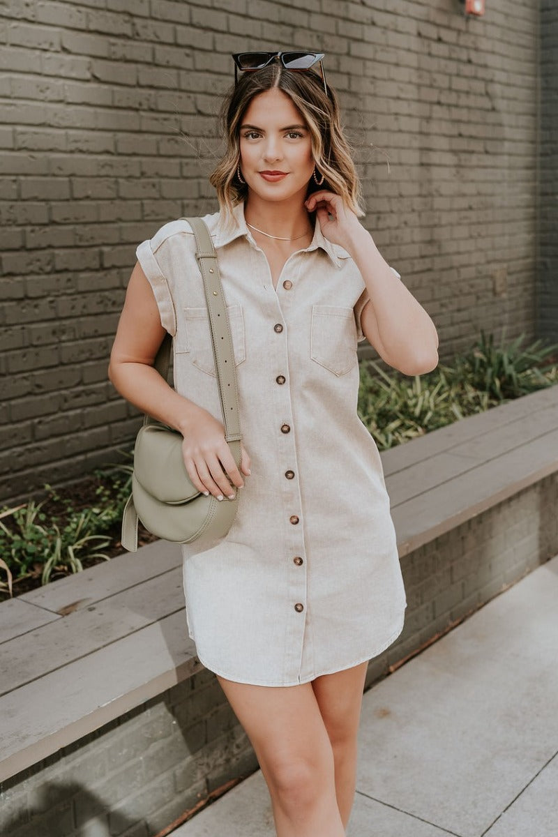 Front view of model wearing the Alexia Taupe Denim Button Up Mini Dress that has taupe washed denim fabric. side pockets, tortoise buttons, front pockets, a collar, mini length, and short sleeves.