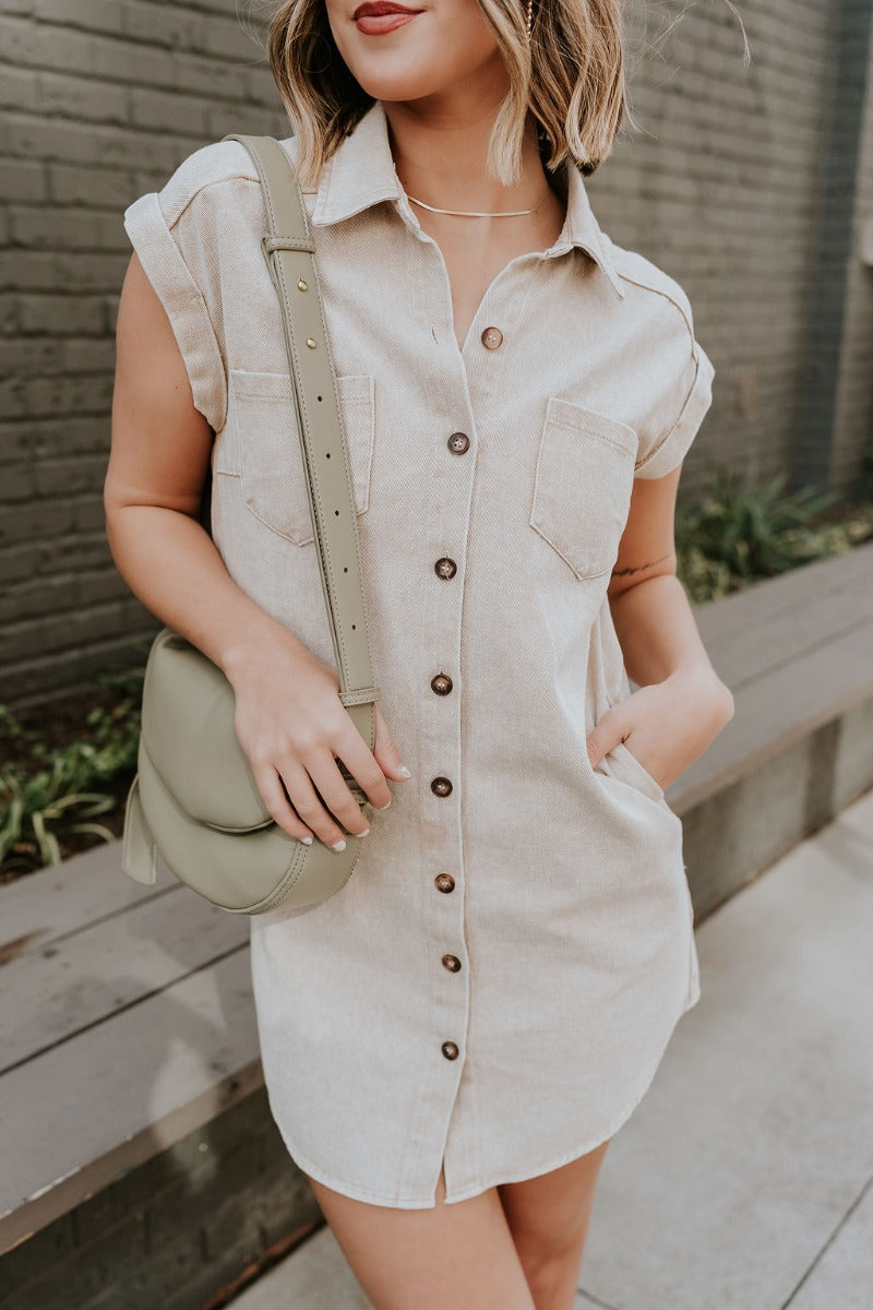 Front view of model wearing the Alexia Taupe Denim Button Up Mini Dress that has taupe washed denim fabric. side pockets, tortoise buttons, front pockets, a collar, mini length, and short sleeves.