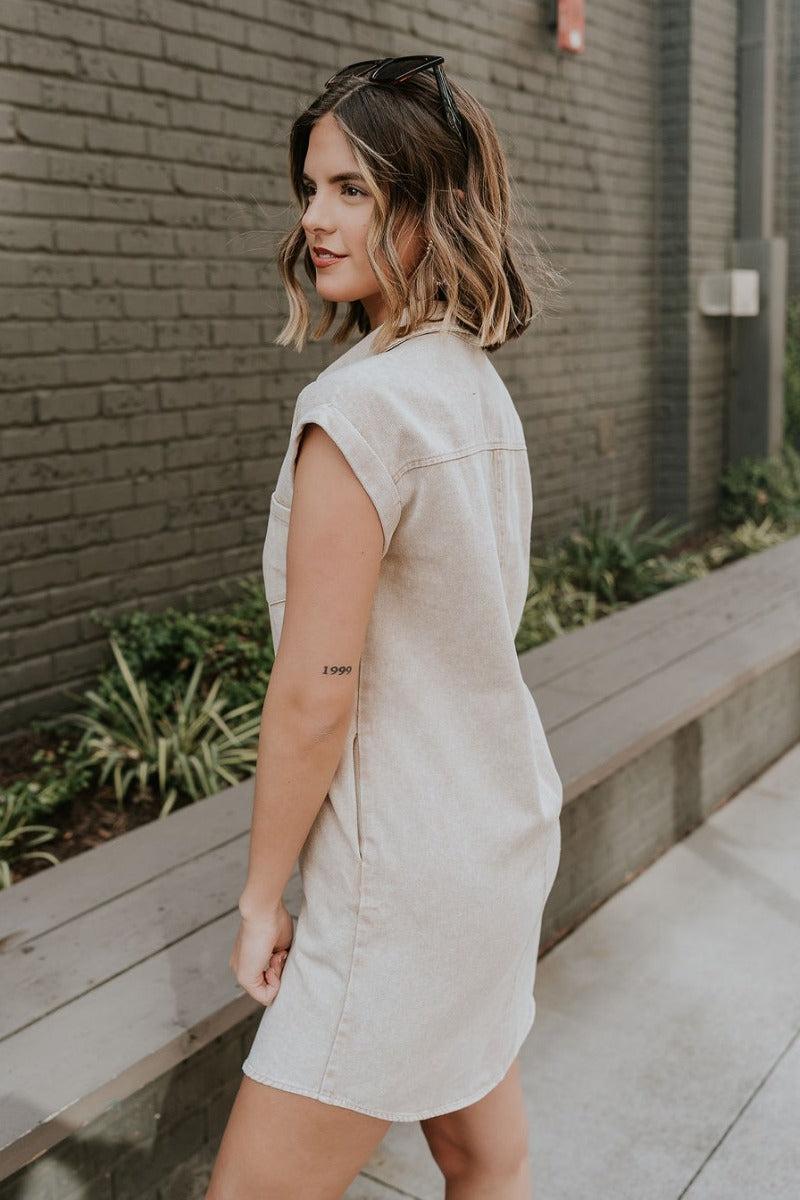 Side view of model wearing the Alexia Taupe Denim Button Up Mini Dress that has taupe washed denim fabric. side pockets, tortoise buttons, front pockets, a collar, mini length, and short sleeves.