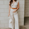 Full body view of model wearing the Nova Beige Sleeveless Belted Jumpsuit which features light beige knit fabric, upper pleated details, wide pant legs, v-neckline, sleeveless, monochrome back zipper with hook closure and an adjustable camel brown belt wi