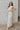 Full body side view of model wearing the Nova Beige Sleeveless Belted Jumpsuit which features light beige knit fabric, upper pleated details, wide pant legs, v-neckline, sleeveless, monochrome back zipper with hook closure and an adjustable camel brown be