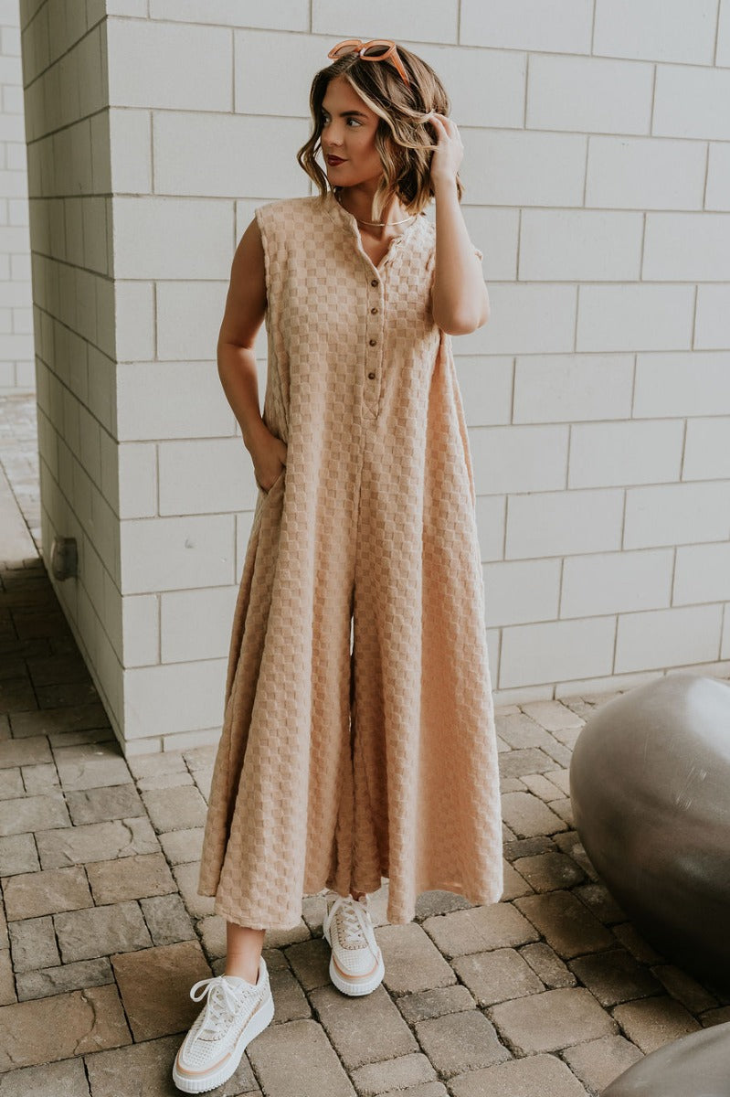 Full body view of model wearing the Mila Taupe Textured Checkered Sleeveless Jumpsuit which features taupe textured fabric, monochrome checkered pattern, two sid pockets, tortoise quarter button up, round neckline, sleeveless and wide pant legs.