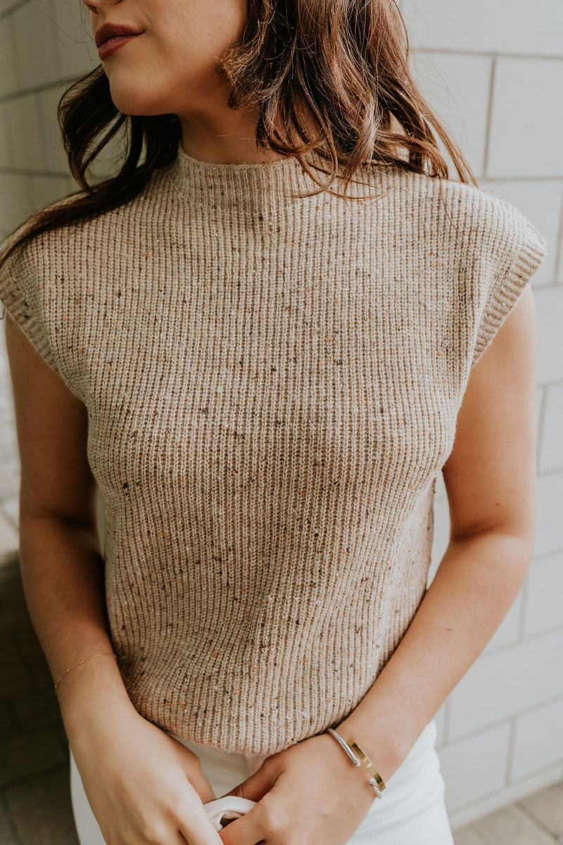 Close up view of model wearing the Clara Beige Multi Sleeveless Sweater which features beige cable knit fabric, dark brown and light brown speckled design, ribbed hem, high neckline and sleeveless.