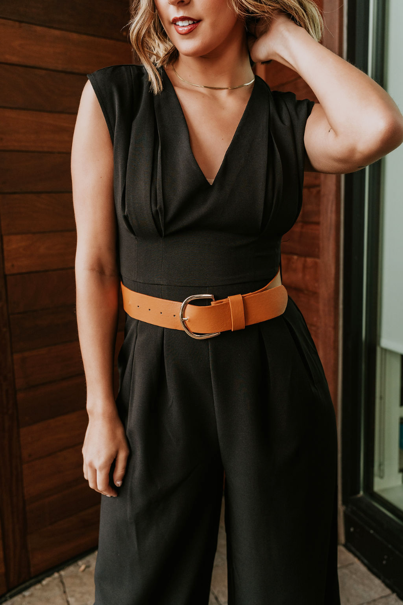Front view of model wearing the Nova Black Sleeveless Belted Jumpsuit which features black knit fabric, upper pleated details, wide pant legs, v-neckline, sleeveless, monochrome back zipper with hook closure and an adjustable camel brown belt with a gold 